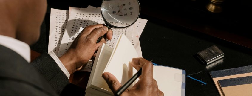 Black private detective seen from behind referencing a notebook as he attempts to decode cryptic text. He holds a magnifying glass in his left hand and a pen in his right. - Visit KinseyInvestigations.com.