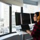 Woman standing at a computer screen display wearing yellow headphones looking out the window at the business district outside - Visit KinseyInvestigations.com.