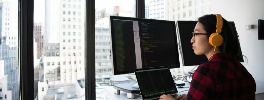 Woman standing at a computer screen display wearing yellow headphones looking out the window at the business district outside - Visit KinseyInvestigations.com.