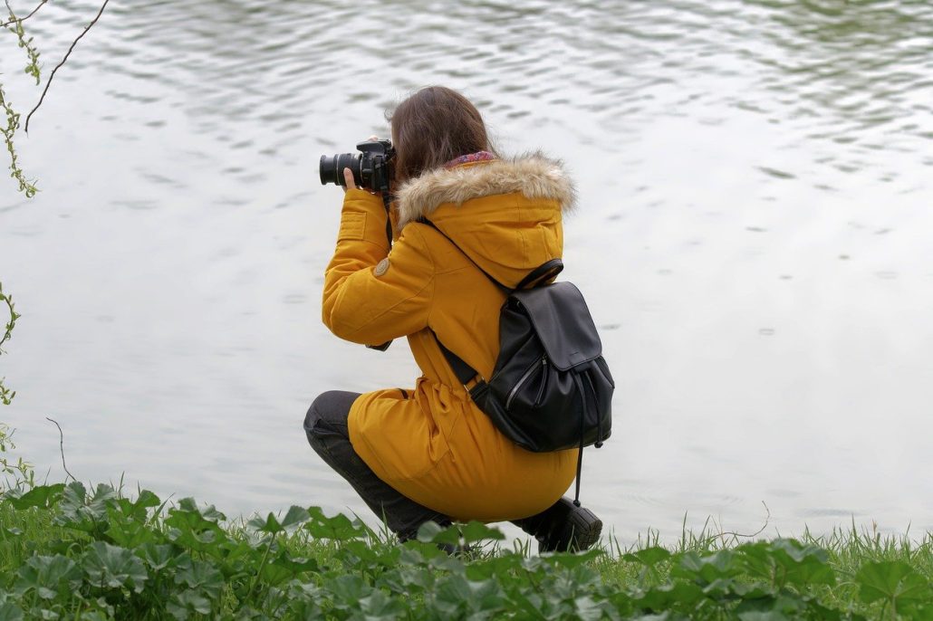 Female photographer in yellow coat with black backpack by waterfront - Visit KinseyInvestigations.com. 