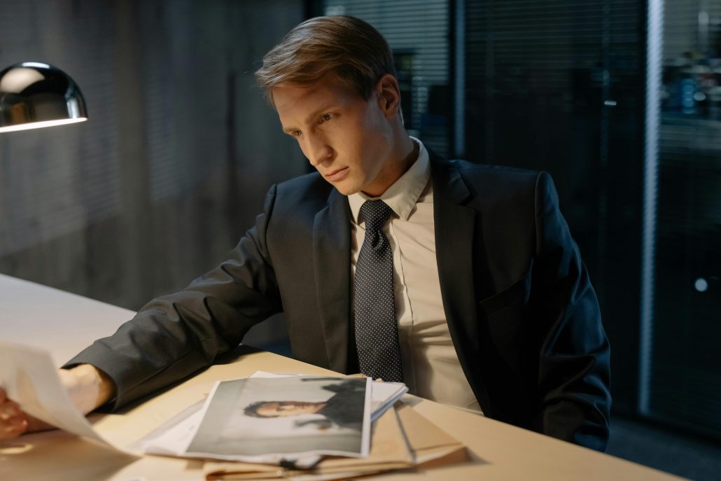 A white man in a suit seated in an office examines printed photos - Visit KinseyInvestigations.com. 