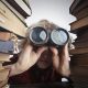 Blonde woman with binoculars peering out from between two stacks of books - Visit KinseyInvestigations.com.