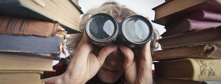 Blonde woman with binoculars peering out from between two stacks of books - Visit KinseyInvestigations.com.