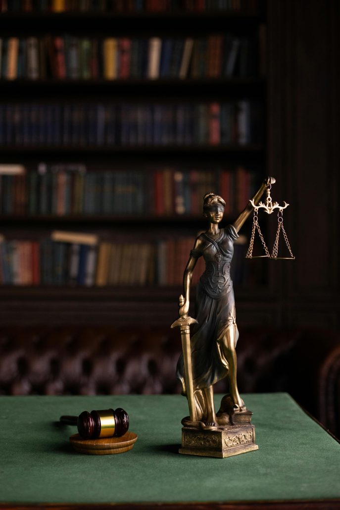 Small statuette of justice with sword in one hand in scales on a desk in an office, books lining shelves in the background - Consult KinseyInvestigations.com.