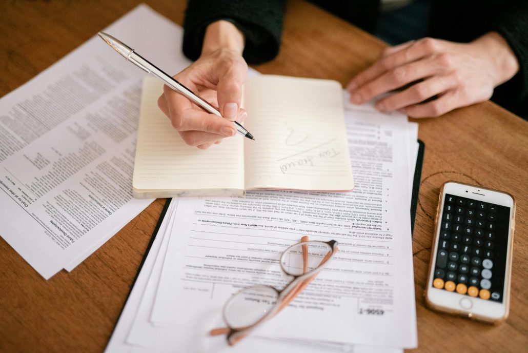 A person with a pen in their hand takes notes on some documents. A pair of glasses rests on the documents. - Visit KinseyInvestigations.com.