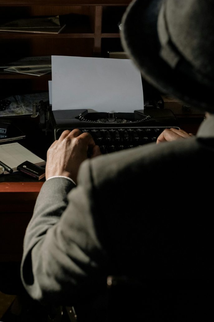 A man in an old-fashioned suit seen from behind sits at a typewriter - Visit KinseyInvestigations.com.