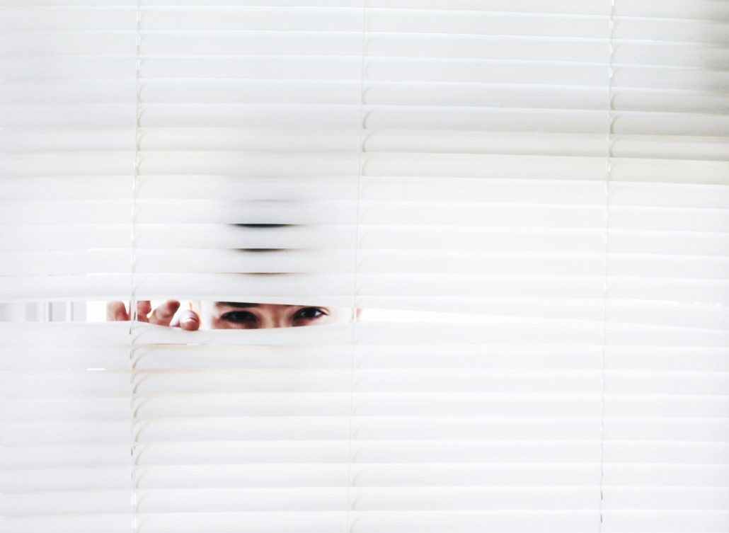 A man's eyes peeking through blinds in a window. 