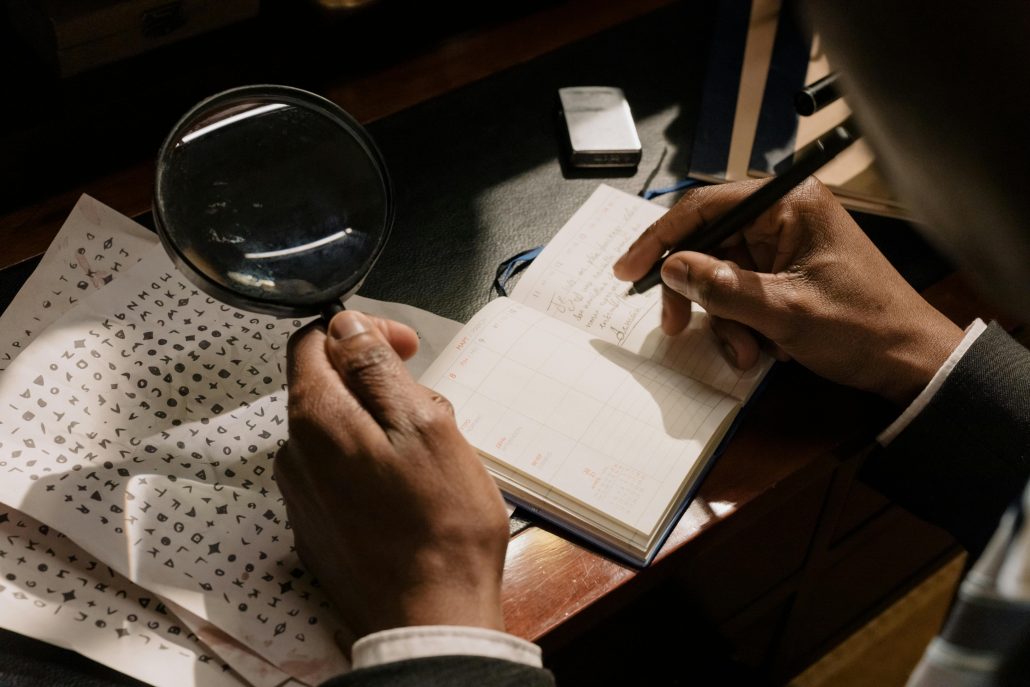 A man in a suit with a magnifying glass in one hand decrypting a code on a notebook - Visit KinseyInvestigations.com. 