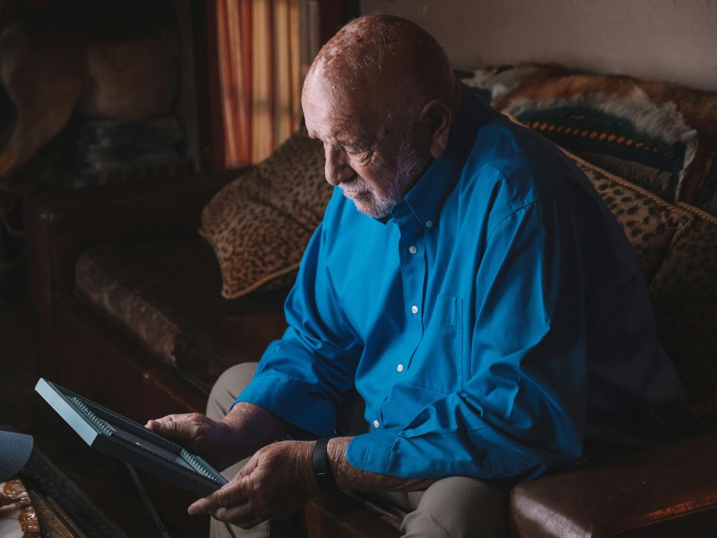 A sad elderly man on a couch looking down at a framed photo - Contact KinseyInvestigations.com.