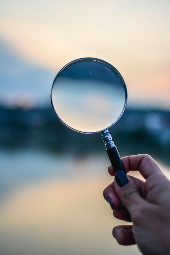 A hand holding up a magnifying glass. The background is indistinct. - Visit KinseyInvestigations.com. 