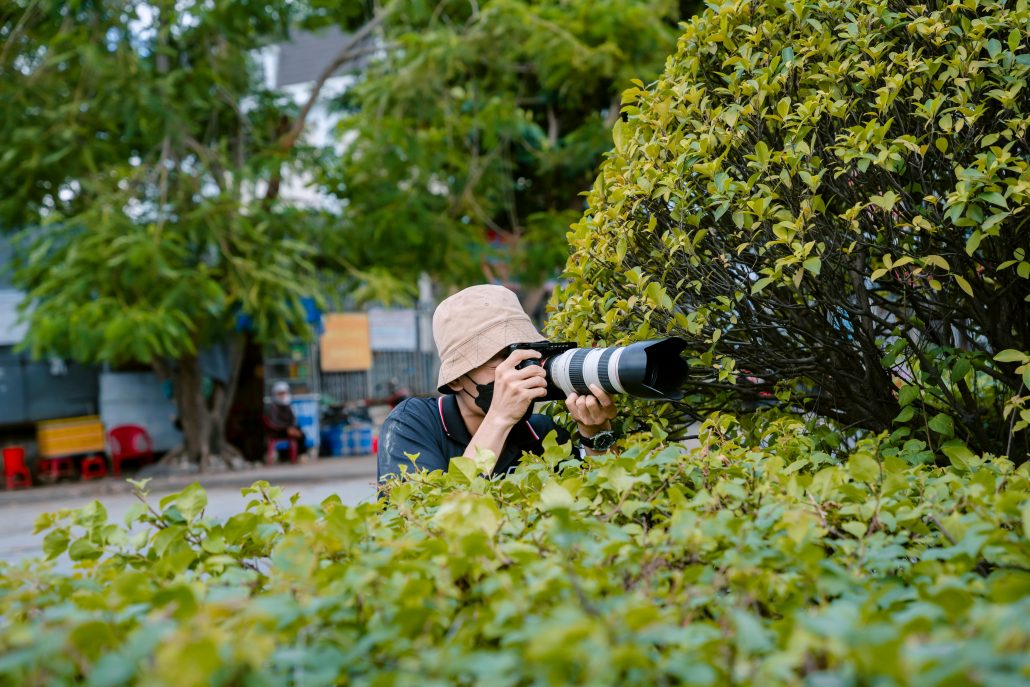 A photographer in a hat and mask behind a bush - Visit KinseyInvestigations.com. 