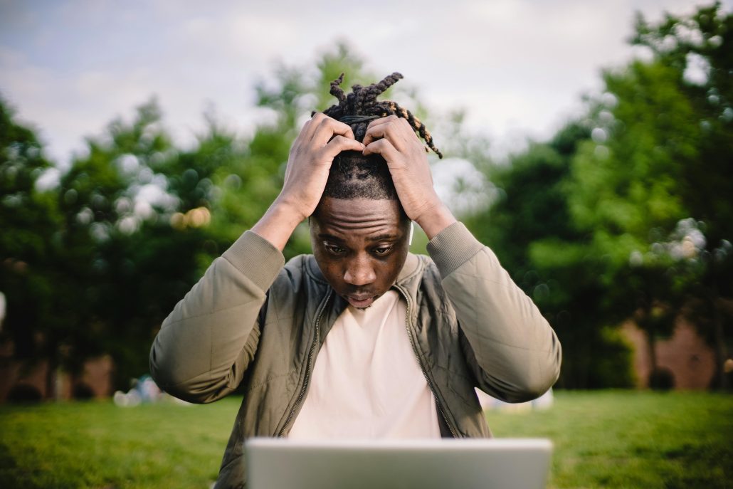 A man with his hands in his hair in frustration sitting in front of a laptop outdoors - Consult KinseyInvestigations.com. 