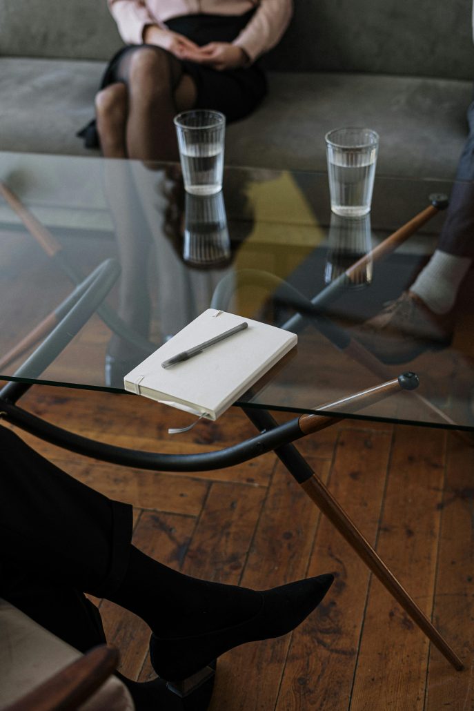 A notebook and pen sit on a glass table. A couple's feet are seen beneath it across from a woman in heels - Visit KinseyInvestigations.com.